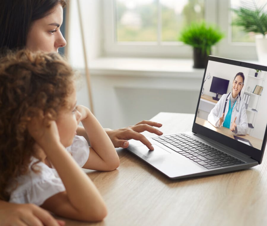 A woman and a young child sit at a table while having a video call on a laptop with a veterinarian. The vet is smiling and wearing a white coat with a stethoscope. The room is well-lit with natural light coming from a window in the background.