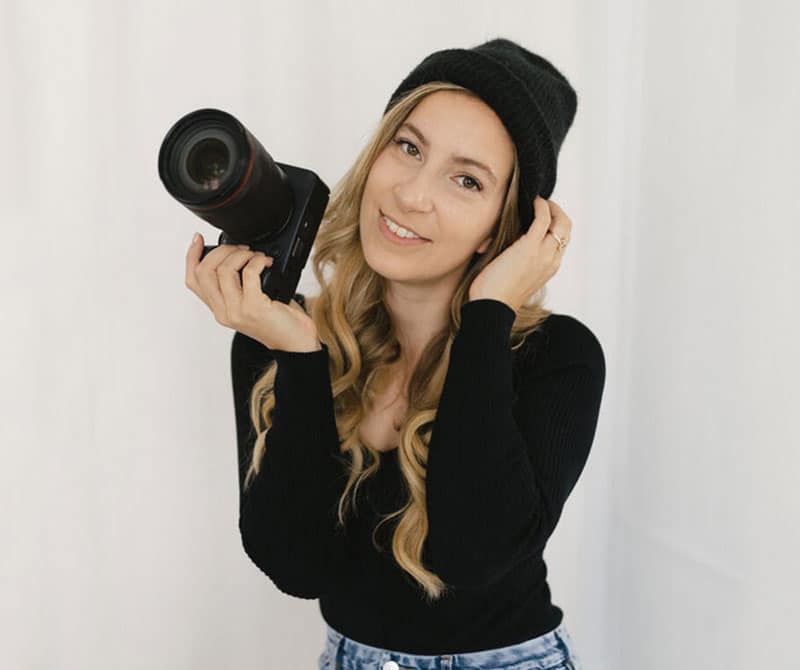 A smiling woman with long, wavy blonde hair holds a camera in one hand and adjusts her black beanie with the other. She is wearing a black top and light blue jeans, standing in front of a plain white background.