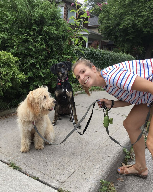 A smiling woman in a striped shirt holds the leashes of two dogs—a shaggy cream-colored one and a black-and-brown one—while standing on a sidewalk with greenery and houses in the background.