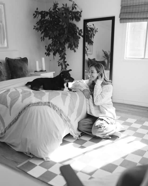 A woman sits on the floor, smiling at a dog lying on a bed in a cozy, sunlit bedroom. A large plant, mirror, and window are in the background. The scene is in black and white.
